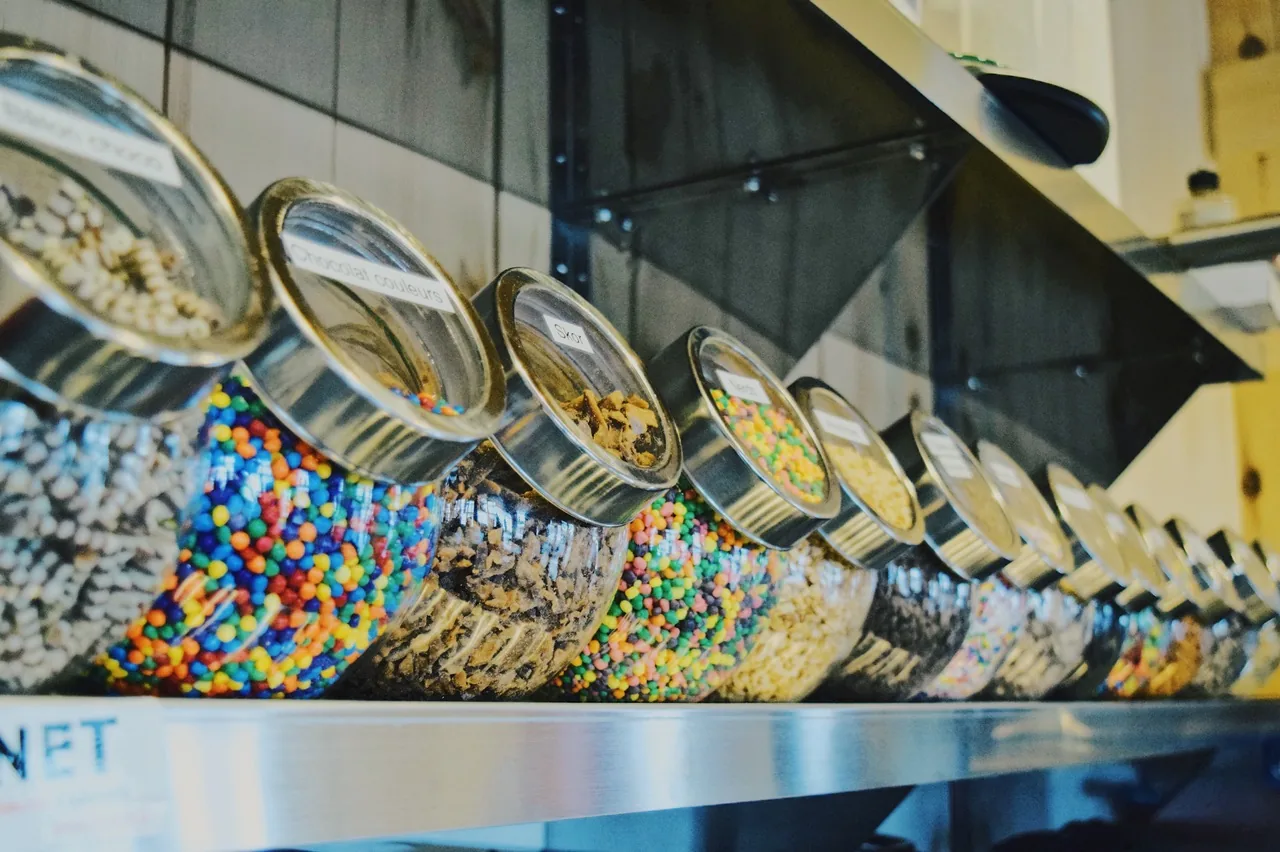 La rangée de pots de garnitures en verre alignés au comptoir : bonbons colorés, Skor, éclats de chocolat.